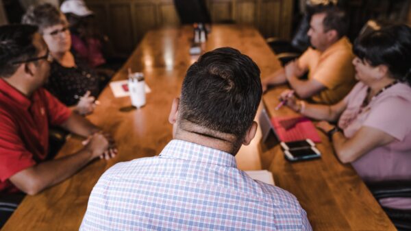 Back of a man's back and head with a table and four other people in the background seemingly at a meeting. The man at the front is wearing a collared white shirt with red and blue striped checkers.