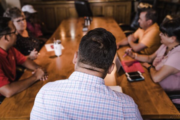 Back of a man's back and head with a table and four other people in the background seemingly at a meeting. The man at the front is wearing a collared white shirt with red and blue striped checkers.