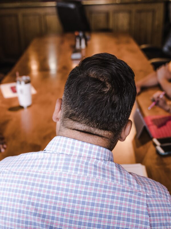 Back of a man's back and head with a table and four other people in the background seemingly at a meeting. The man at the front is wearing a collared white shirt with red and blue striped checkers.