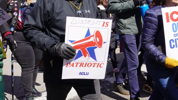 Photo from a protest of a man wearing a black jacket and gloves holding a sign with a megaphone and a star graphic, with the text "dissent is patriotic" and the ACLU logo.