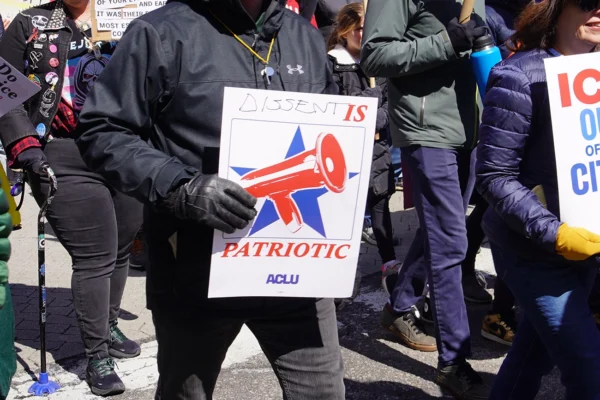 Photo from a protest of a man wearing a black jacket and gloves holding a sign with a megaphone and a star graphic, with the text "dissent is patriotic" and the ACLU logo.