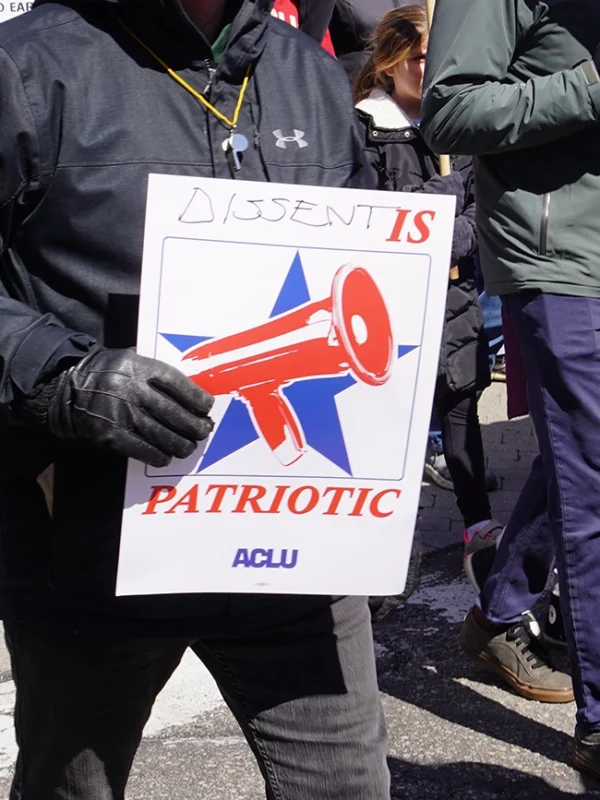 Photo from a protest of a man wearing a black jacket and gloves holding a sign with a megaphone and a star graphic, with the text "dissent is patriotic" and the ACLU logo.