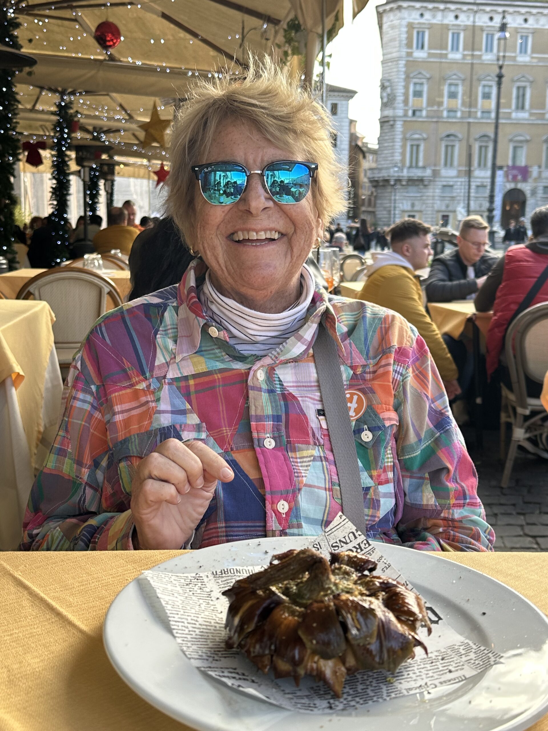 White woman with short blonde hair and blue sunglasses in a colorful jacket and white turtleneck shirt pointing at a plate of food.